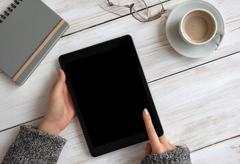Photo of a person holding an iPad with a black screen on a white desk, next to a coffee cup and a notebook, in a top-down view. Web banner with copyspace for text or a logo on the right side 