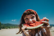 © qunica.com - A cheerful young woman bites into a watermelon slice on a bright summer day, capturing the joy of nature and outdoor fun near a scenic beach and lake environment.