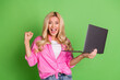 © deagreez - Excited young woman celebrating success while holding a laptop, wearing a casual pink shirt, standing against a vibrant green background
