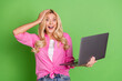 © deagreez - Young woman surprised while using laptop, expressing joyful astonishment, casual attire, blonde hairstyle, green background