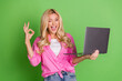© deagreez - Cheerful young woman showcasing positivity while holding a laptop and making a gesture against a vivid green studio background