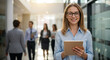 © schiers_images - A woman is standing in a bright office hallway, holding a tablet and smiling at the camera, with blurred colleagues walking in the background.