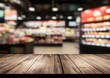 © multasam - Wooden table top in front of a blurred supermarket interior  Display shelves filled with various products, lights, and general store ambiance are visible in the background
