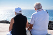 © luciano - Rear view of caucasian senior pensioner couple sitting face the sea enjoying freedom and vacation, two elderly people in retirement stay together in good harmony. Horizon over water