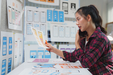 Wall Mural - young asian woman working as a ux ui designer, engaging in a phone conversation while discussing the