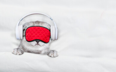 Leinwandbilder - Cute kitten wearing  headphones and mask for sleep listening music on a bed at home. Top down view. Empty space for text