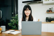 © Nuttapong punna - Asian woman working remotely on a laptop and tablet in a cozy kitchen workspace. Perfect for education, freelance
