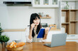 © Nuttapong punna - Asian woman working remotely on a laptop and tablet in a cozy kitchen workspace. Perfect for education, freelance