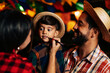 © kleberpicui - Family celebrating Brazilian Festa Junina. A child in plaid shirt and straw hat gets a mustache drawn by his mother. Colorful flags and lights in the background