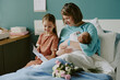 © AnnaStills - Mother sitting in hospital bed, holding newborn, while daughter sits nearby with a card. Room filled with happiness, flowers, and a blue blanket