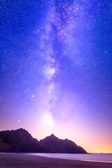 Wall Mural - Vibrant purple and blue night sky with the Milky Way galaxy arching over a dark silhouette of coastal mountains and a sandy beach