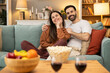 © Dejan - A joyful couple cuddles together on a couch, surrounded by snacks, as they enjoy a movie night in a cozy living room setting.