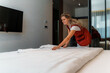© DusanJelicic - Smiling hotel maid arranging crisp white sheets on a neatly made bed in a modern hotel room, creating a clean and inviting atmosphere for guests to enjoy