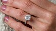 © Ant - A hand with light pink nails wears a sparkling diamond engagement ring featuring a round-cut stone and a pave band.
