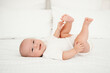 © Any Grant - a laughing baby in a white bodysuit on his back on the bed at home smiling, a portrait of a baby on the bed, a small child holding or playing with his legs, a place for text.