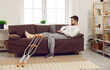 © Studio Romantic - Handicapped young man lying at home on sofa in rehabilitation with his crutches and working on a laptop. Brunet guy in rehab after injury with injured leg typing on computer or chatting with friends.