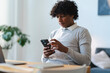 © Studio Marmellata - Focused young Arab man using smartphone at home. He is sitting at a desk with a laptop and notebook. He is wearing a gray sweater and white collared shirt.