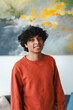 © Studio Marmellata - Portrait of a smiling young Arab man. He is wearing a casual orange sweater and standing indoors with artwork behind him.