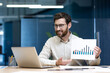 © Tetiana - A young male businessman works in an office, sits at a desk and talks on a video call, shows graphs and documents to the camera