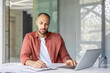 © Liubomir - A focused man, dressed in a red shirt, is working diligently at a desk, surrounded by documents and a laptop, in a modern office.