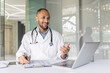 © Liubomir - A doctor wearing a lab coat smiles while talking during a video conference from his office. He's using a laptop and gesturing.