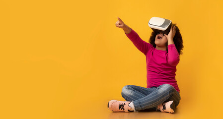 Leinwandbilder - Little Afro Girl Wearing VR Headset, Experiencing Virtual Reality And Pointing Away While Sitting On Floor Over Yellow Background In Studio, Panorama