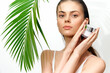 © VICHIZH - Young woman with natural beauty holding a skincare cream near a palm leaf, showcasing freshness and vitality in a bright, tropical setting.