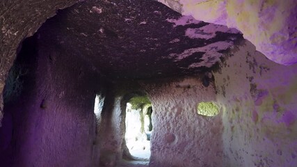 Wall Mural - Ancient rock-carved tunnel hallway with textured stone walls, purple light reflections, and small natural openings allowing sunlight into the space.