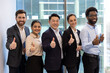 © Liubomir - A diverse team of business professionals give a thumbs up while looking at the camera, happy with success.