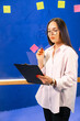 © Vadim - Young woman brainstorming in an office. A young woman with glasses is deep in thought while reviewing notes at a creative workspace adorned with sticky notes.