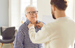 © Studio Romantic - Adult son helping his senior father to put on his clothes. Young man buttons up the shirt of his happy, cheerful, smiling old dad. Family, older age, care concept