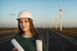 © hayoshka - Female engineer in hard hat with two plans by curved road, looking at wind field view.