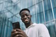 © marimalina - Man smiles confidently while using smartphone outside a modern glass building in bright daylight