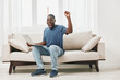 © SHOTPRIME STUDIO - Happy African man celebrating success while using a laptop on a beige sofa, bright room with natural light, casual wear, modern interior