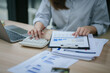 © PSG - Young asian businesswoman holding pen and reading financial documents, smiling and working at office desk with computer showing graphs, startup business concept