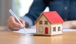 © Picstings - Person signing documents with a toy house model nearby, symbolizing real estate decisions.