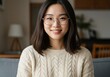 © SimpleDesignStudio - A young adult Asian woman with dark hair and round glasses smiles warmly at the camera, wearing a cozy beige cable-knit sweater indoors in a bright, comfortable setting.