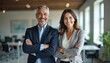 © Viktor - Smiling business man and woman stand arms crossed in office. Happy mature pro leaders posing together. Hispanic man, European woman in formal suits looking camera.