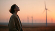 © Lucianastudio - Serenity and sustainable energy a woman embraces nature's beauty with wind turbines at sunrise