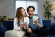 © amnaj - Excited couple holding and showing money while sitting on sofa at home, celebrating financial success, lottery win or profitable investment
