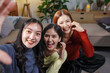 © amnaj - Three cheerful young women sitting on the floor in a cozy living room, capturing joyful moments together while taking a fun selfie