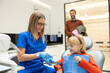© Dejan - A young girl in a dental chair smiles while a dentist prepares for a checkup, showcasing a friendly dental environment.