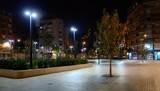Nighttime urban plaza with illuminated buildings and trees.  Well-lit paved area, low-level landscaping, and modern streetlights