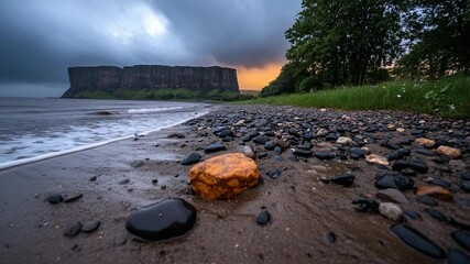 Aufkleber - Coastal landscape at dusk with an orange and black rocks on the sand foreground a large mountain and sunset glow in the background