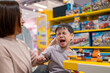 © Parichat - Stubborn child refusing to leave toy store while building with colorful bricks, making mother tired