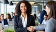 © S-DESIGN - Two business woman shaking hands in a modern office environment, symbolizing partnership and agreement. The woman in front smiles and exudes professionalism, surrounded by a collaborative team.