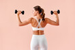 © HBS - Athletic, toned woman turned with her back to the camera, showcasing her beautiful muscular back and arms while posing with dumbbells she is lifting, standing on a soft pink background in a studio.