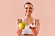 © HBS - Smiling athletic woman in white fitness clothing holding an apple and a dessert, choosing between healthy and sweet food on a soft peach studio background.