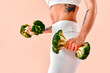 © HBS - An unrecognizable image of a fit and athletic girl in white fitness clothing with a tattoo on her stomach, holding broccoli like dumbbells. Healthy eating and fitness.
