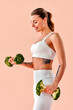 © HBS - A fit, athletic girl in white fitness clothing is lifting broccoli in her hands as if they were dumbbells, against a soft pink background. Creative advertisement, concept of fitness and healthy eating